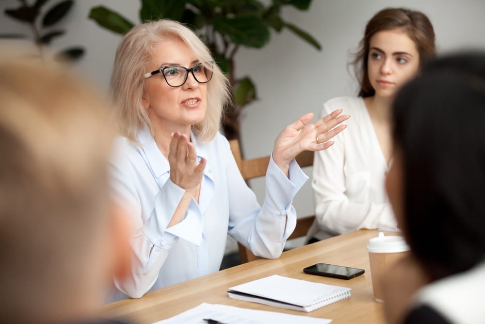 A confident older woman with glasses gestures while speaking during a group meeting, surrounded by colleagues and discussion materials—illustrating a collaborative approach to conflict resolution - Conflict Coaching