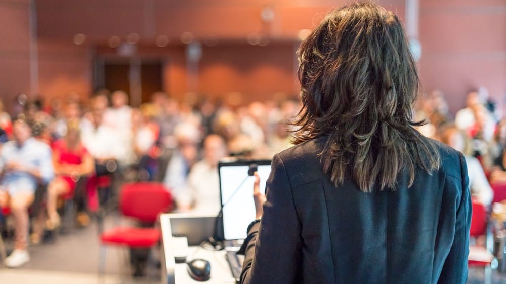 A professional woman stands at a podium facing a seated audience in a conference room, delivering a presentation or training session on effective interpersonal strategies - Conflict Coaching